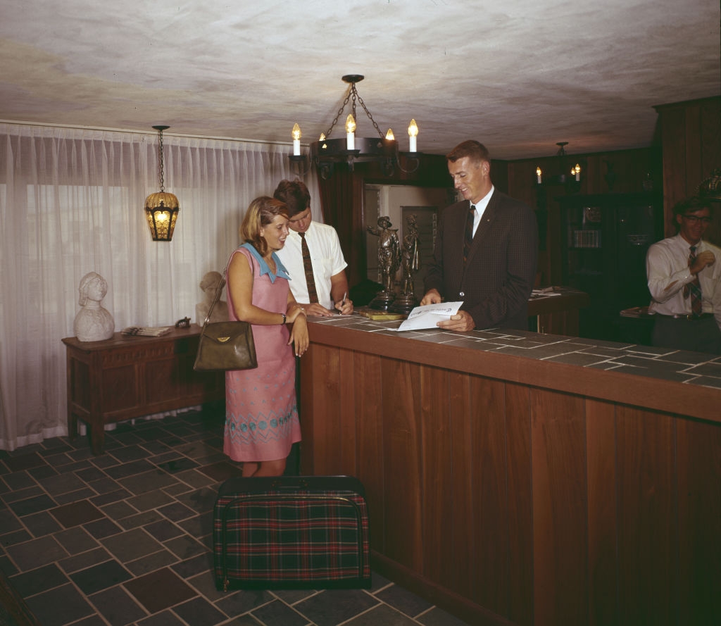 A couple checks into a hotel at the front desk while a receptionist assists them. A plaid suitcase sits on the floor