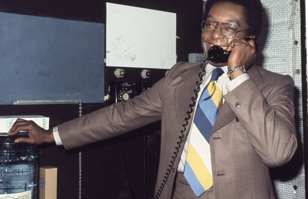 A man in a suit and a striped tie is smiling while talking on a corded phone, standing next to a water cooler. The image is from an article categorized as Rewind