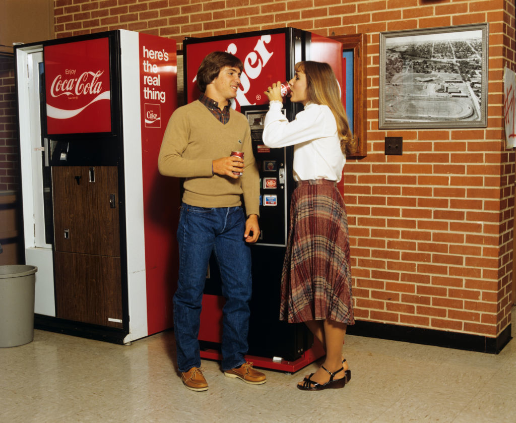 Two people in front of a vintage Coca-Cola vending machine, both holding soda cans. One is wearing a sweater and jeans, the other a white shirt and plaid skirt