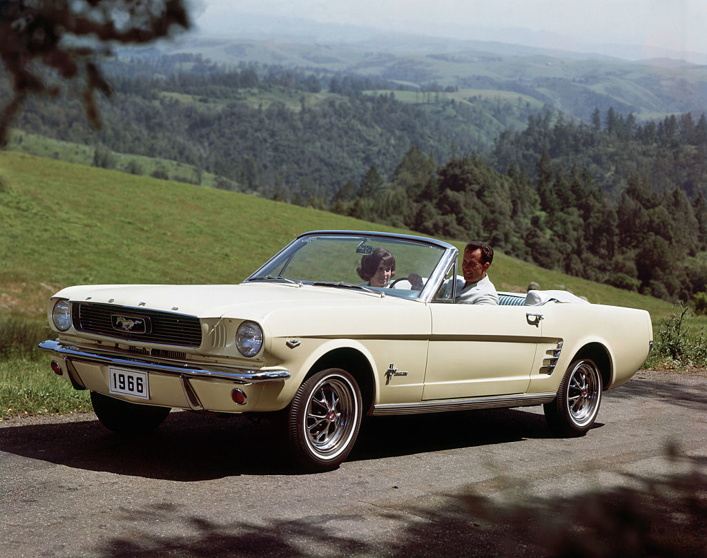 A 1966 Ford Mustang convertible with a man and a woman driving in a scenic countryside setting