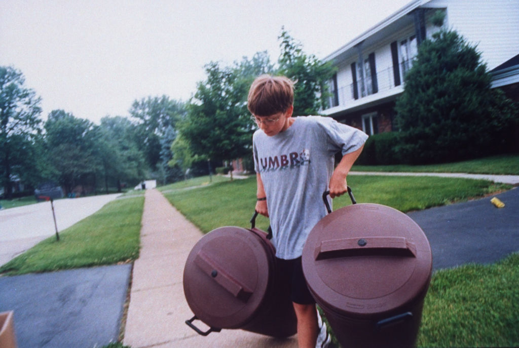 A young boy takes out the trash in a suburban neighborhood, carrying two garbage bins along a sidewalk
