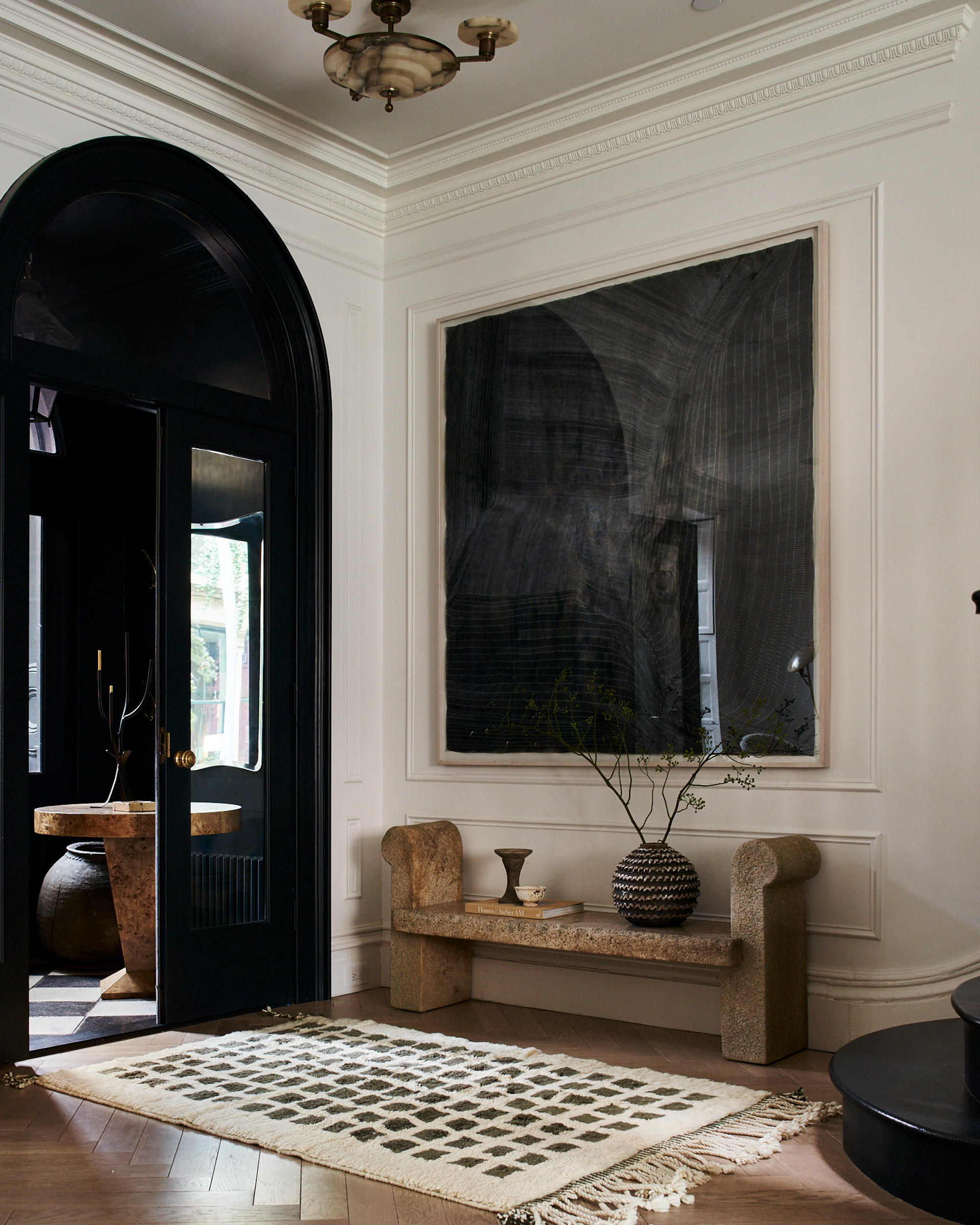 A large entryway in Athena Calderone's house. It is white with a large black painting on the wall and a black, arched doorway at the entry. A black and white checkered rug is on the ground, and there is a stone bench under the painting.