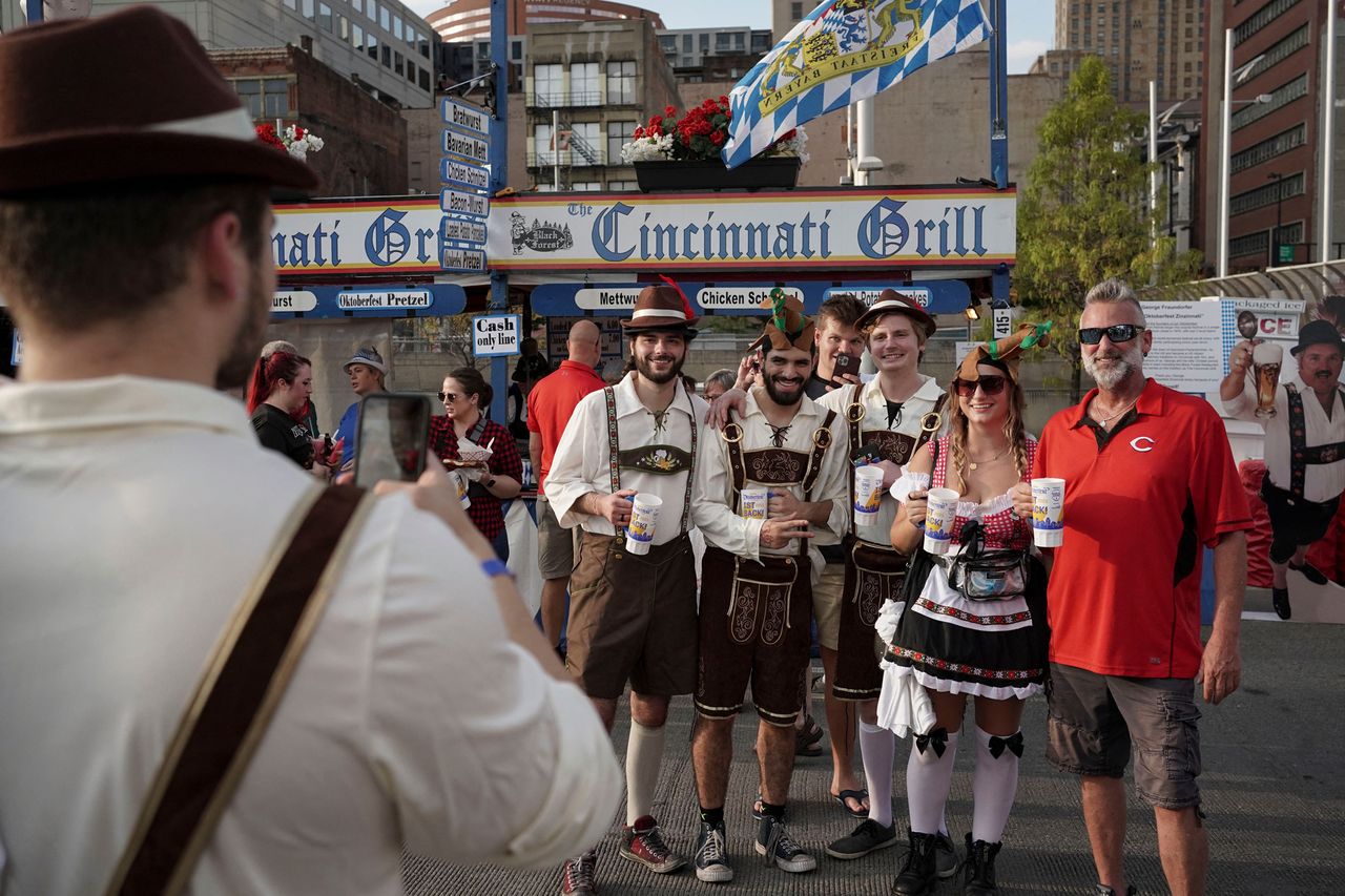Group of six people posing outdoors some wearing German outfits and holding drinks a Cincinnatithemed background is visible