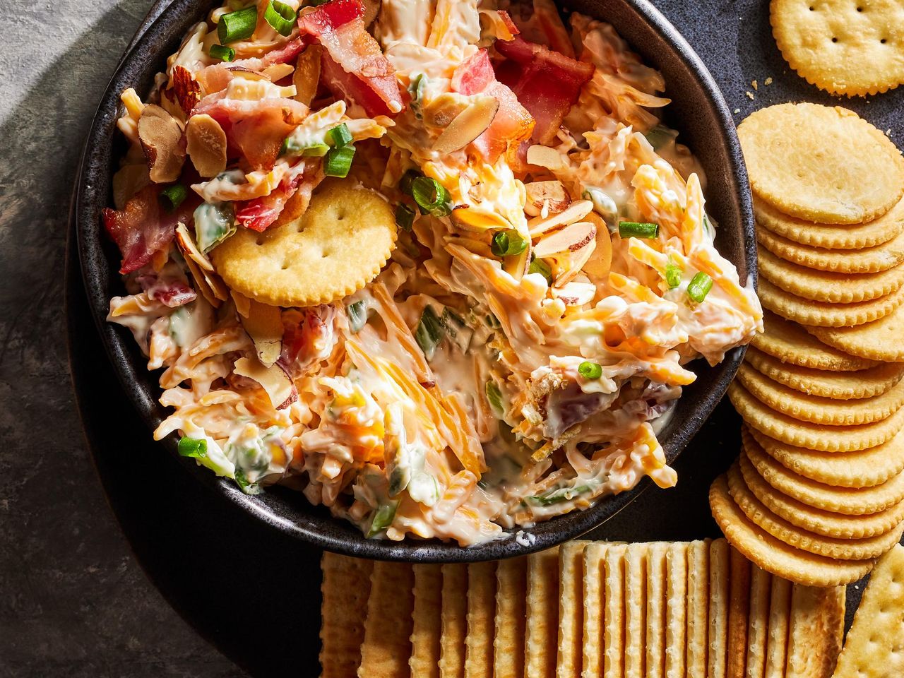 A dish containing a creamy dip garnished with bacon and green onions, surrounded by crackers