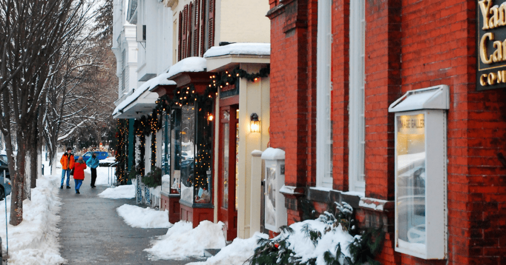 a few people bundled up in winter coats walk down a sidewalk past snow drifts and businesses decorated for christmas in stockbridge ma
