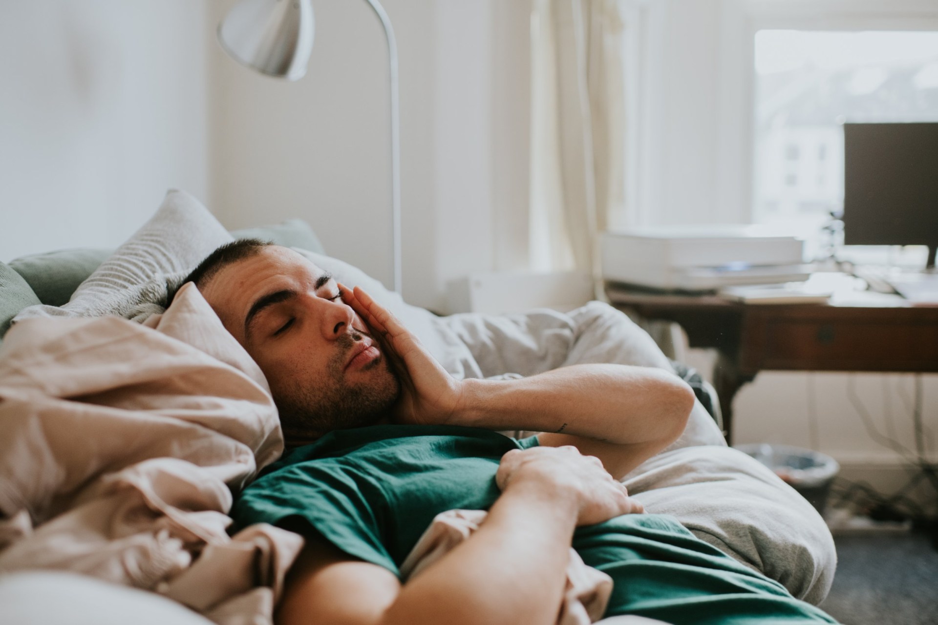 A man has just woken up in a domestic environment, either a living room or a bedroom. He is cozy in duvets and pillows. He rubs his eyes and face sleepily as he comes to.