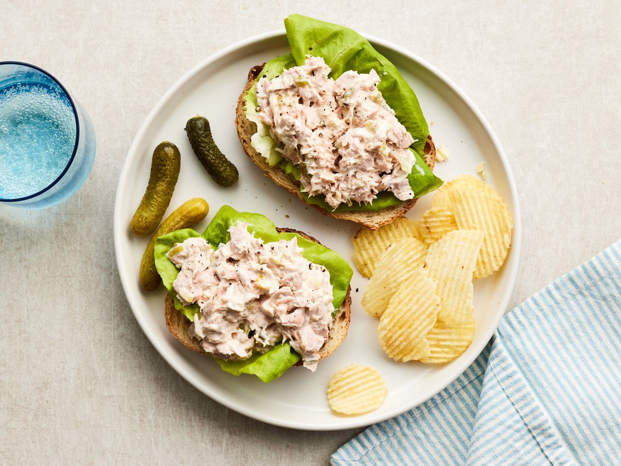 overhead shot of two open face tuna sandwiches on a white plate with gherkins and potato chips