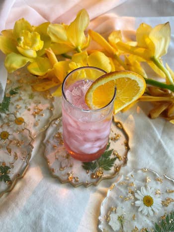 Iced pink drink with an orange slice garnish on floral coasters, surrounded by yellow flowers, creating a refreshing and elegant scene