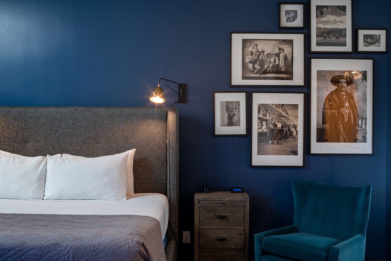 A neatly made hotel bed beside a gallery wall of framed art and a chair