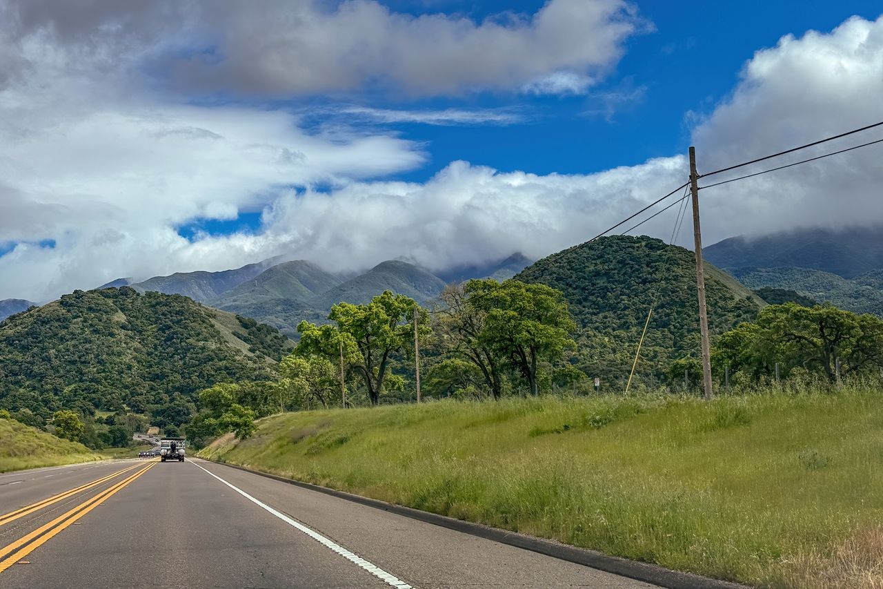 A countryside road heading toward a mountainous area with some clouds and green hills visible