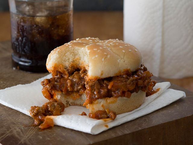 close up view of a Sloppy Joe Sandwich on top of a paper towel, with a glass with soda and a roll of paper towels in the background