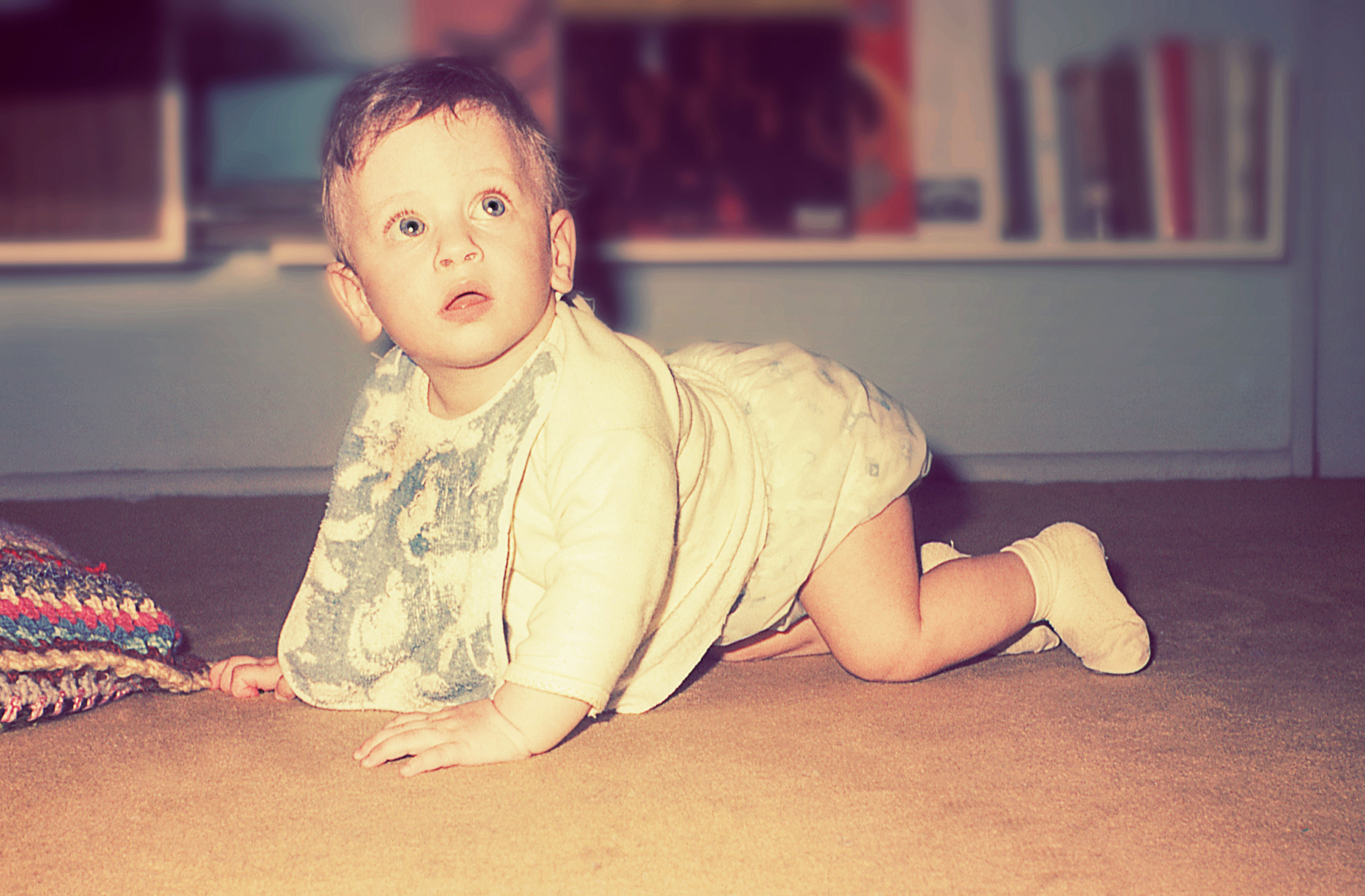 A baby in a bib is crawling on carpet in a cozy room, looking upwards curiously