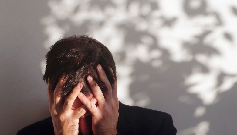 Person with head in hands, surrounded by shadowy leaf patterns on a wall, conveys stress or contemplation