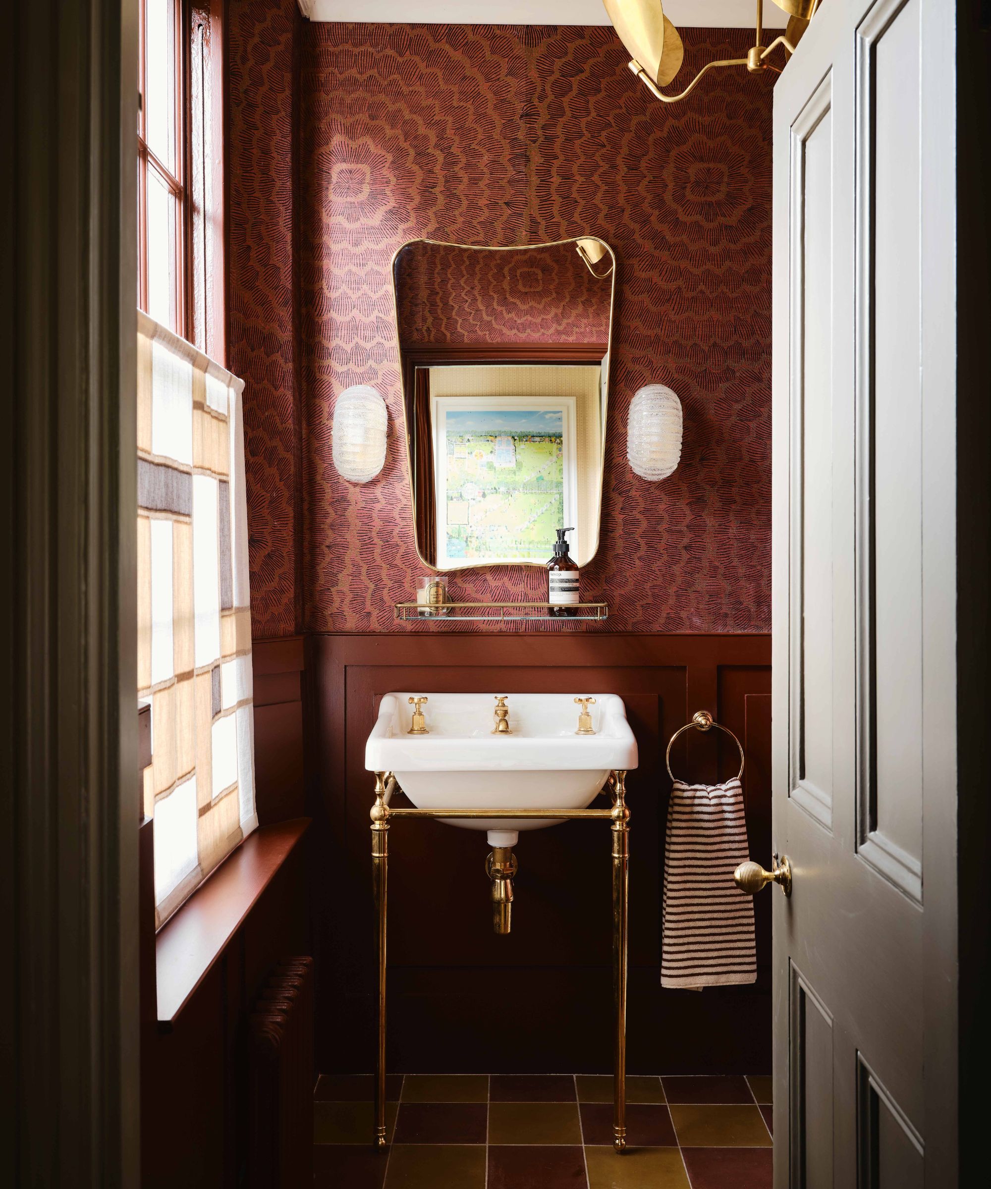 dark burgundy red powder bathroom with panelled walls and wallpaper above, with a mid century mirror, vintage wall sconces, and a traditional sink on a brass stand
