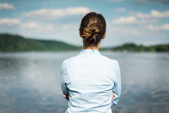 Person with hair in a bun stands by a calm lake, arms crossed, looking at the distant landscape under a partly cloudy sky