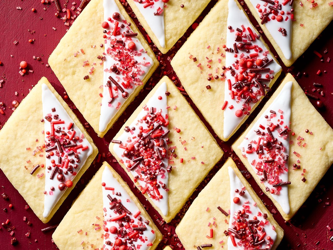 Diamondshaped sugar cookies with icing and red sprinkles arranged in a grid on a red background