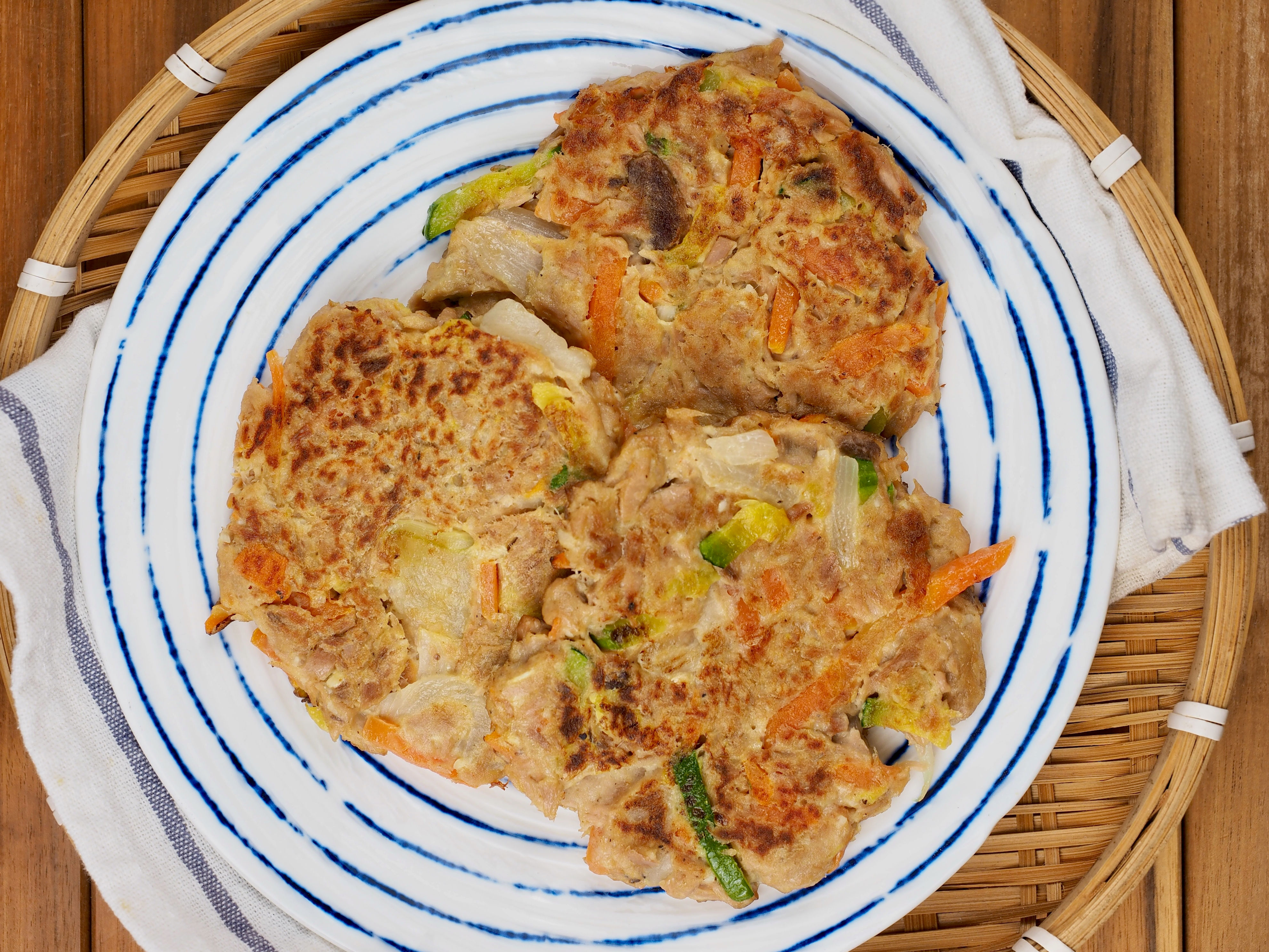 Two vegetable pancakes on a white plate with blue stripes, placed on a wicker tray. Contains visible pieces of carrots and green onions
