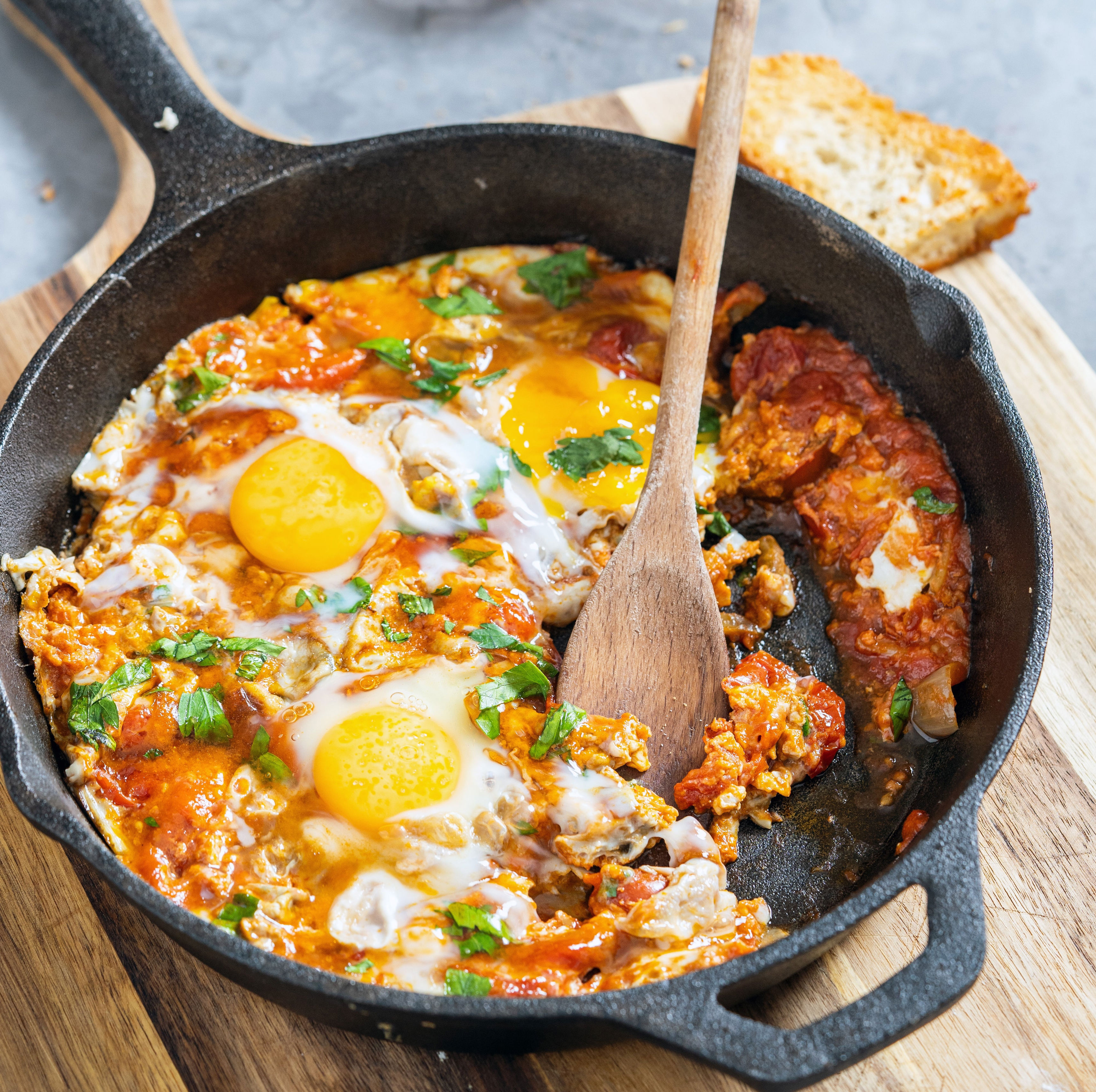 Cast-iron skillet with shakshuka: poached eggs, tomato sauce, herbs. Wooden spoon and toasted bread beside