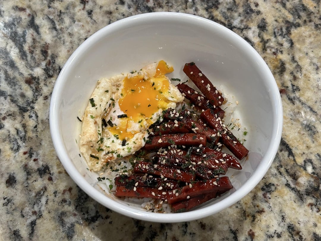 Rice bowl with a fried egg, sliced sausage, and sprinkled herbs in a white bowl on a granite countertop