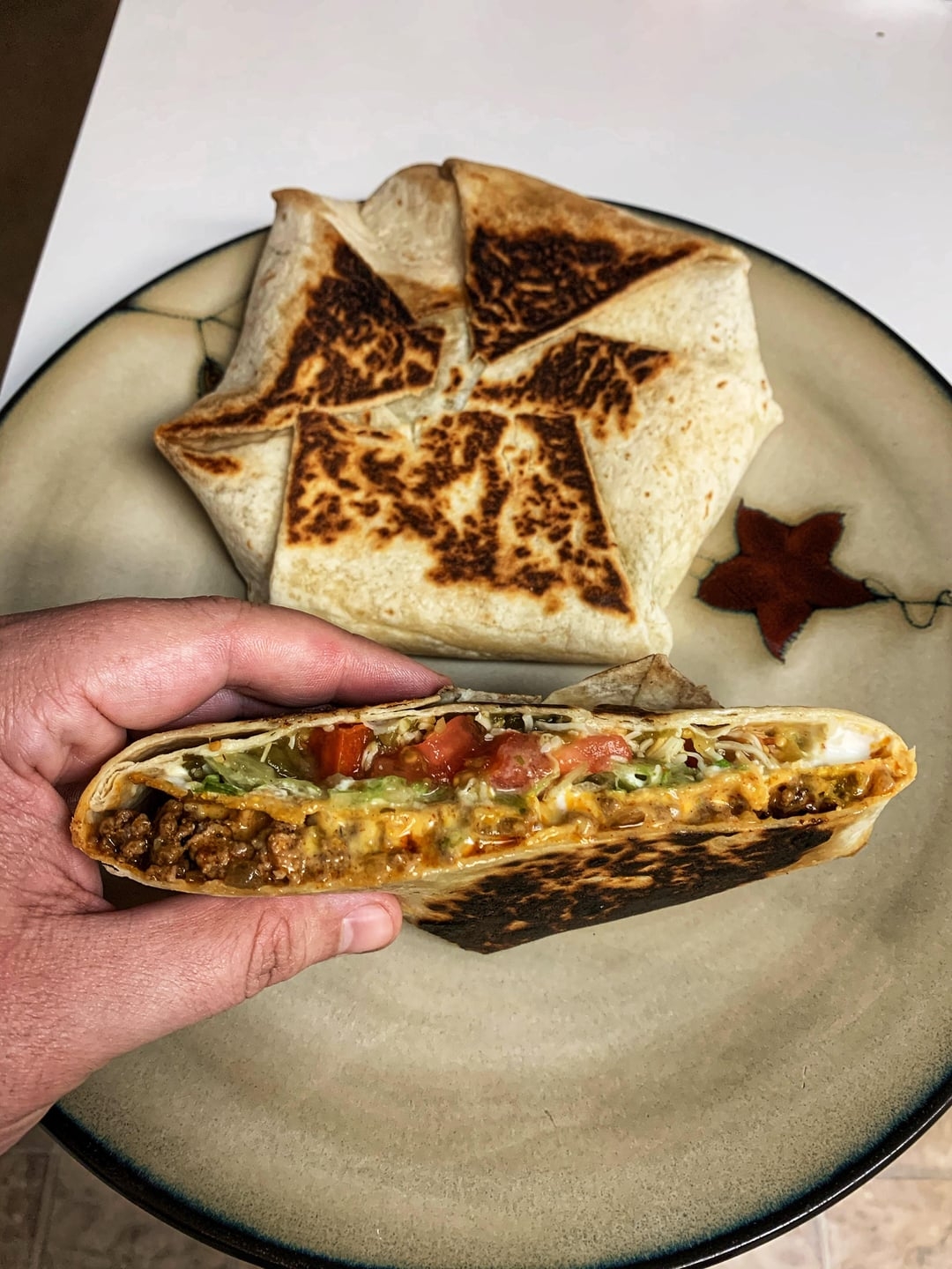 A hand holds a grilled wrap filled with ground beef, cheese, lettuce, and tomatoes, with a similar uncut wrap on a plate in the background