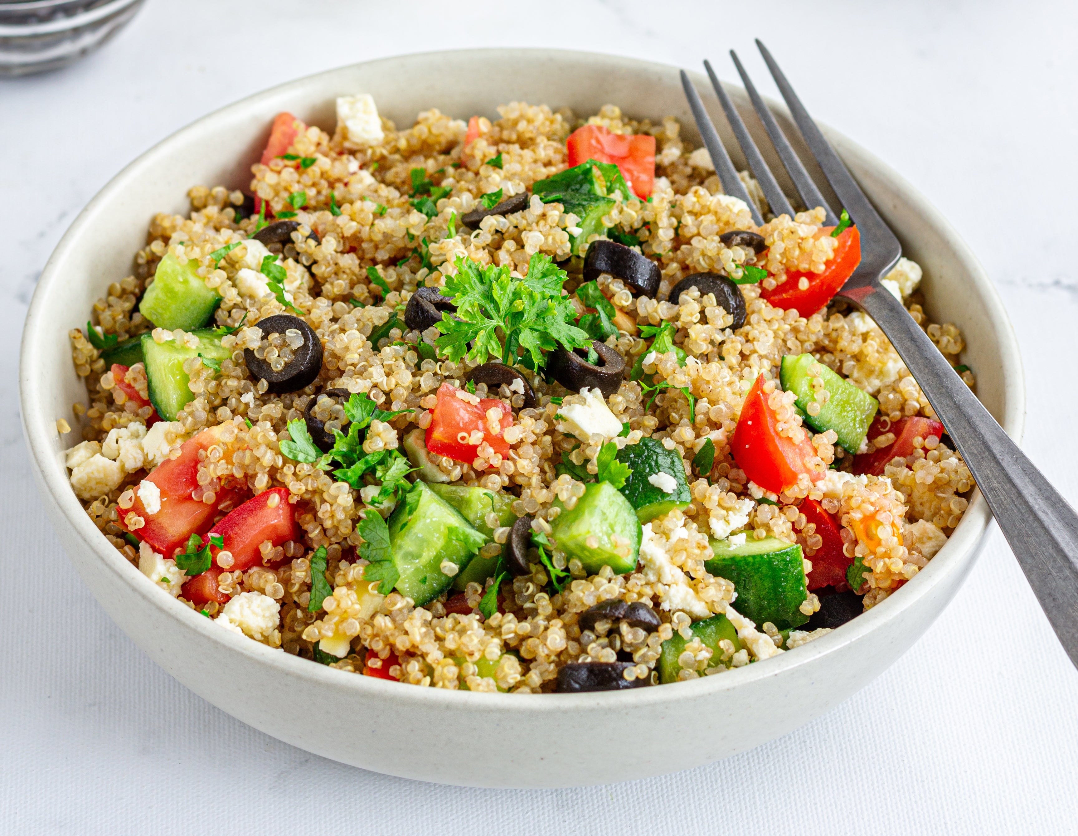 Bowl of quinoa salad with tomatoes, cucumbers, olives, and parsley. Bowl of tomatoes and olive oil in the background