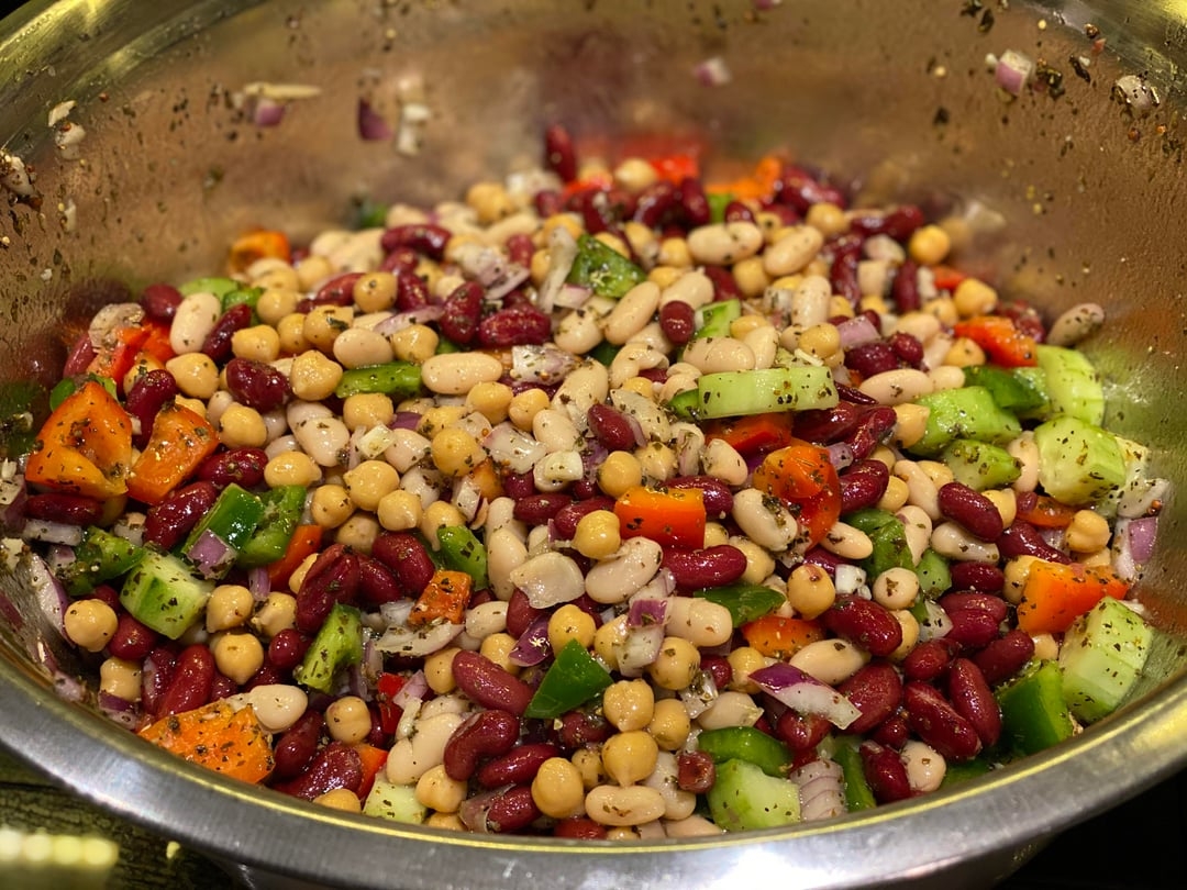A bowl of mixed bean salad with chickpeas, kidney beans, bell peppers, cucumbers, and red onions, topped with a sprinkle of herbs