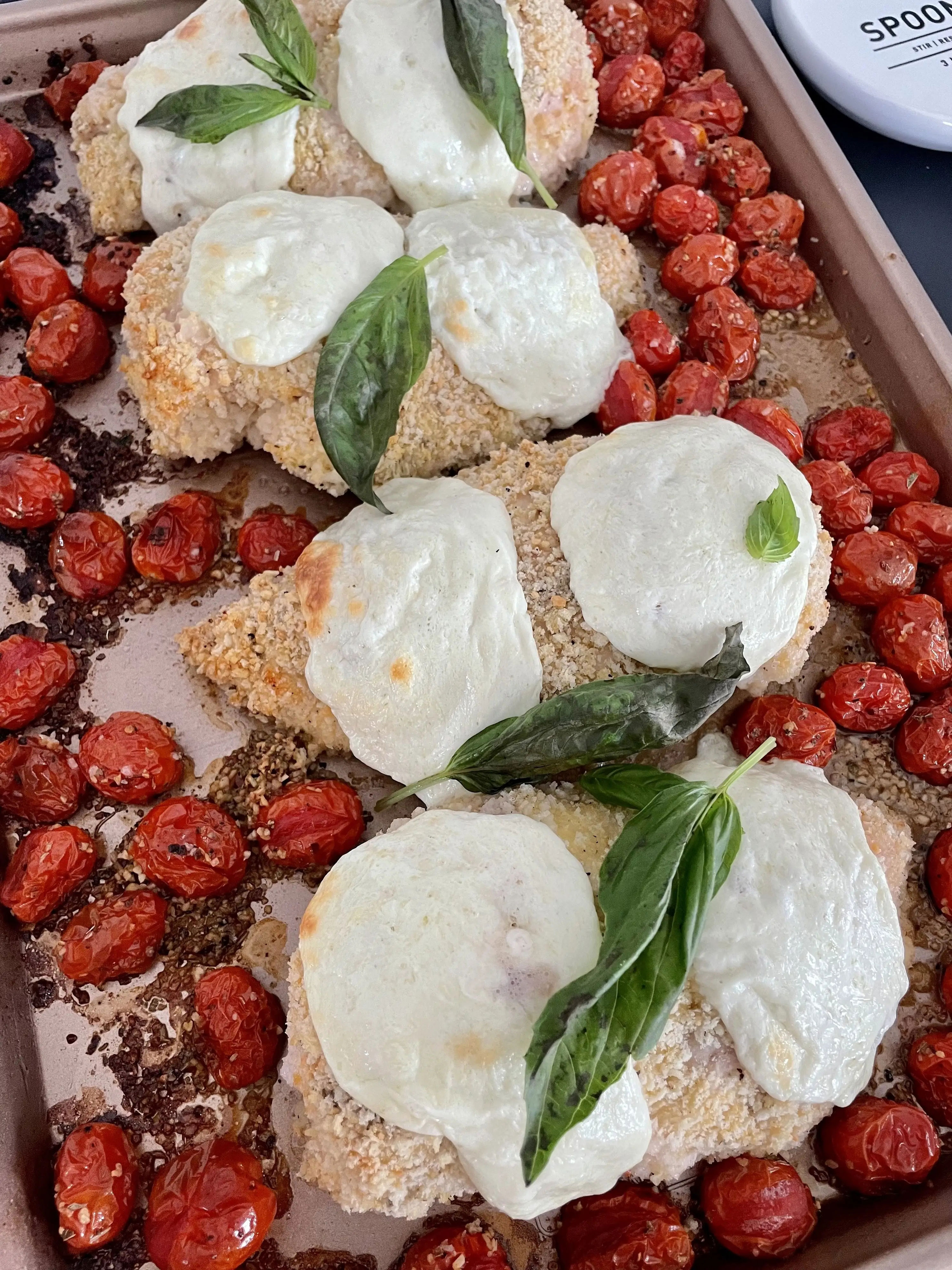 Baked chicken breasts with melted mozzarella, fresh basil, and roasted cherry tomatoes on a sheet pan