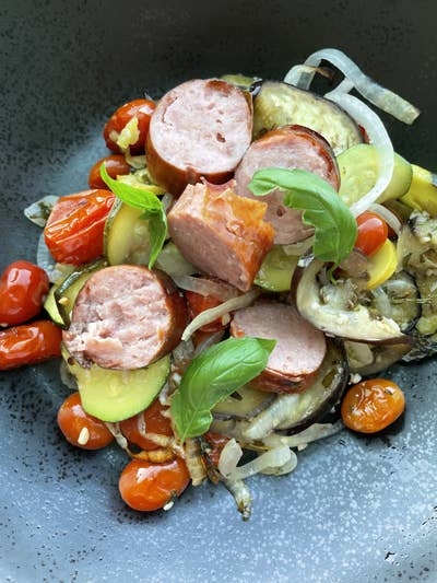 Grilled sausages with sliced vegetables, cherry tomatoes, and basil leaves artfully arranged on a dark plate
