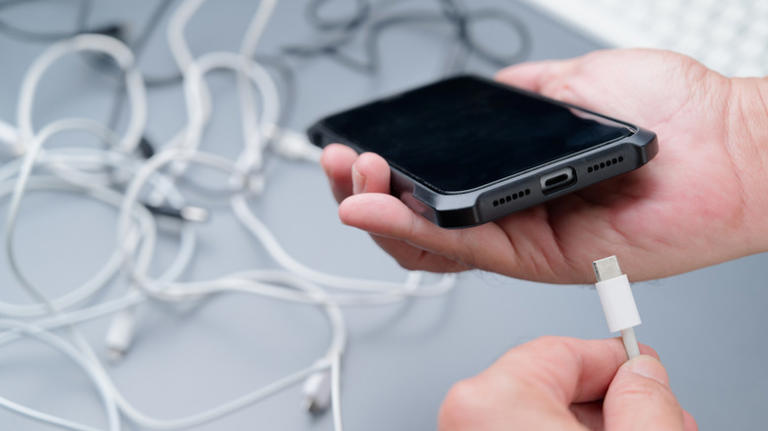 person holding smartphone with usb cable below it, cables behind