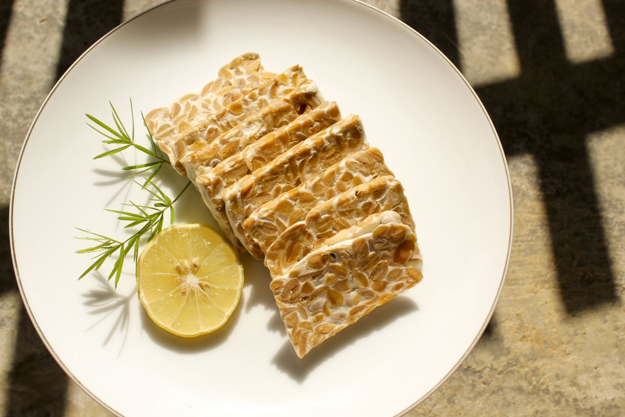 Sliced tempeh on a white plate with a lemon slice and a sprig of herbs.