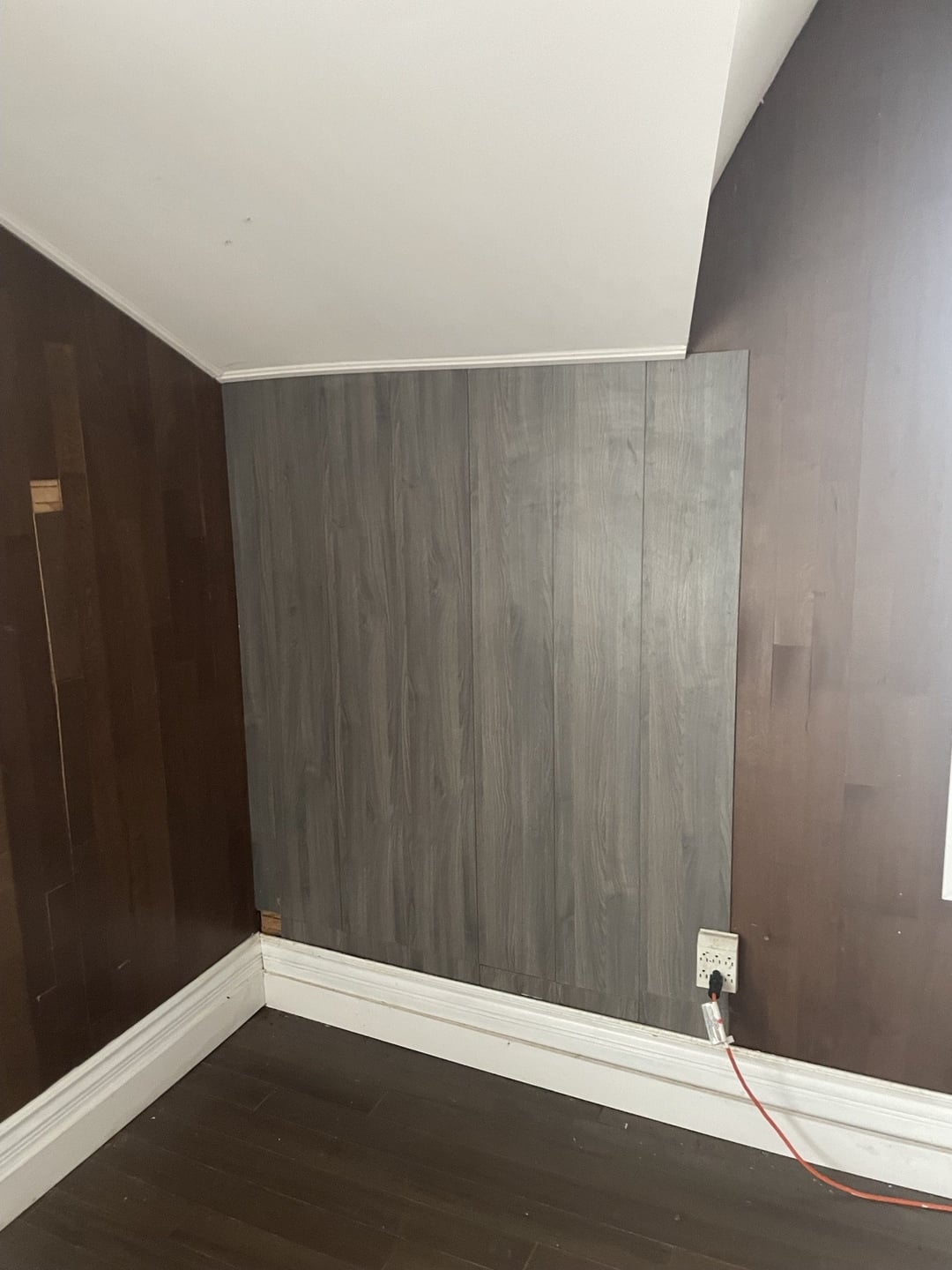 Room corner with mismatched wood panels on walls. A cable is plugged into an outlet near the floor