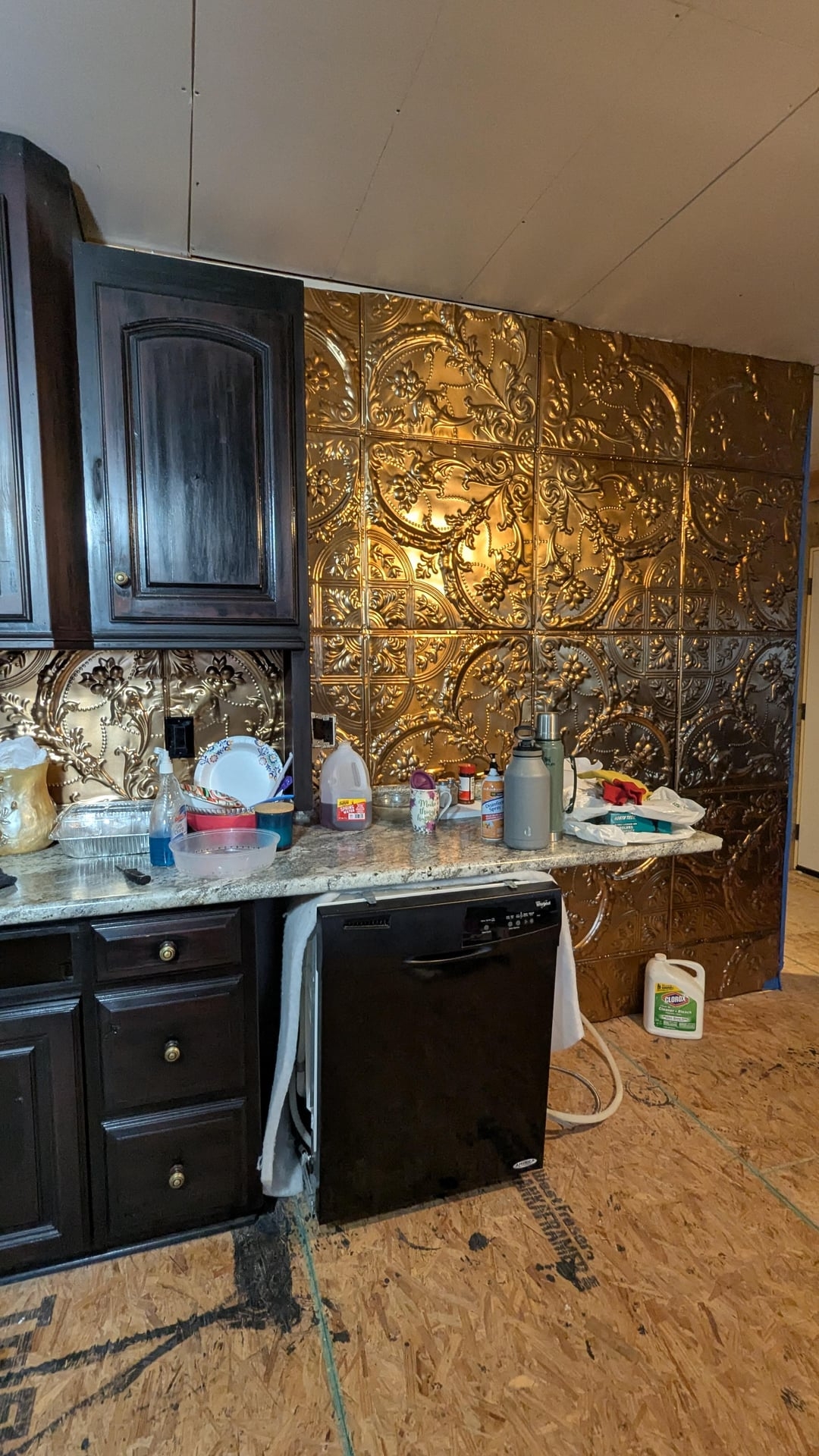 Kitchen with decorative gold-patterned backsplash, dark cabinets, countertop cluttered with dishes and groceries, unfinished wooden floor