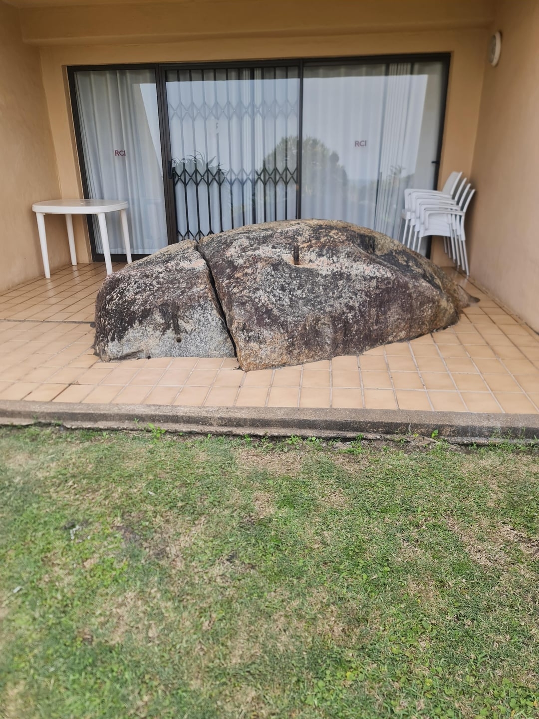 Large rock embedded in a tiled patio area outside a sliding glass door, with a table and stacked chairs nearby on a lawn