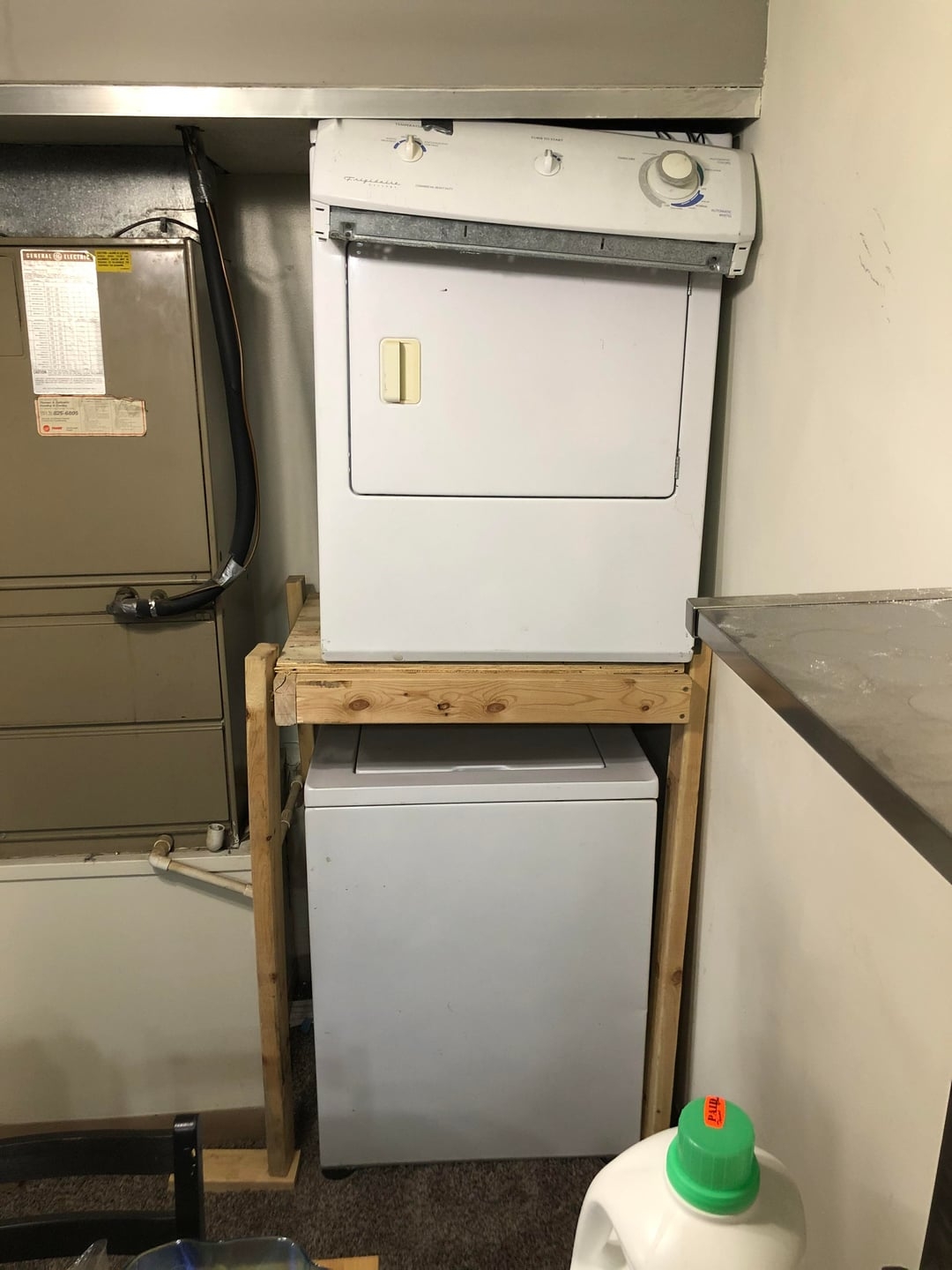 Homemade wooden frame holds a washing machine below and a dryer above, saving space in a utility area. Nearby detergent suggests recent use