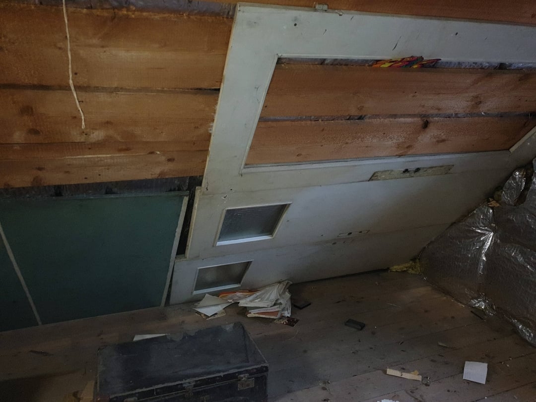Attic space with an angled wooden ceiling, old door against wall, scattered papers, and a partially visible toolbox in a cluttered room