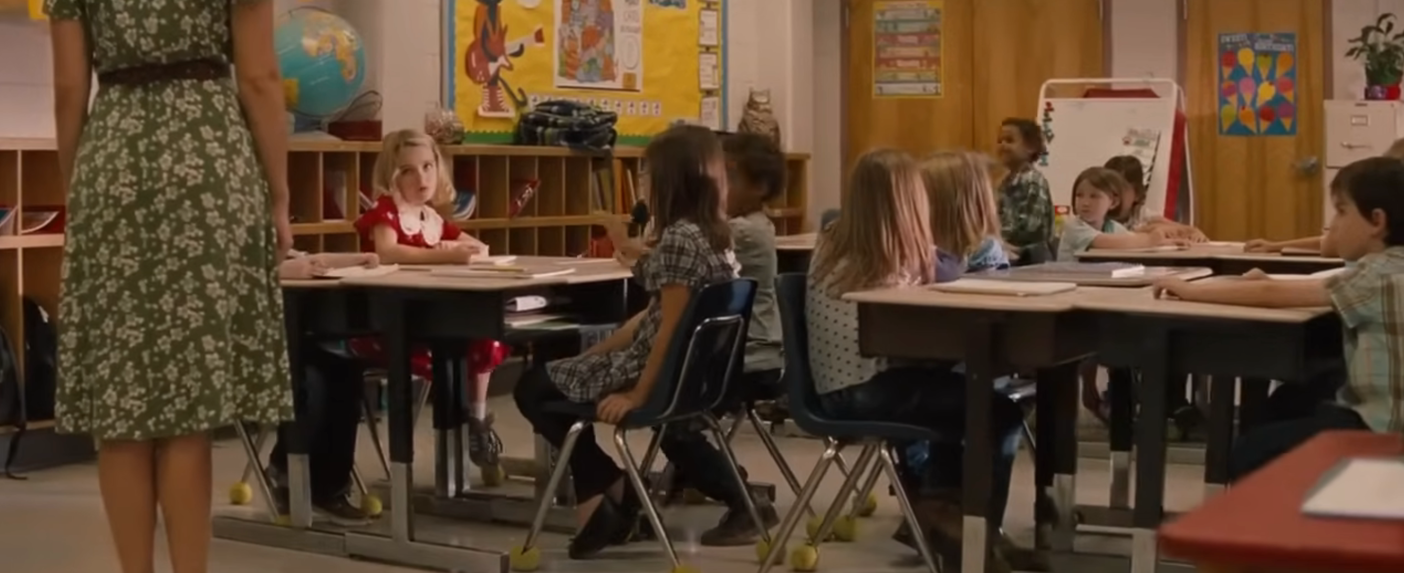 A classroom with young children seated at desks, a teacher stands in front of them facing the class. The room is decorated with educational posters
