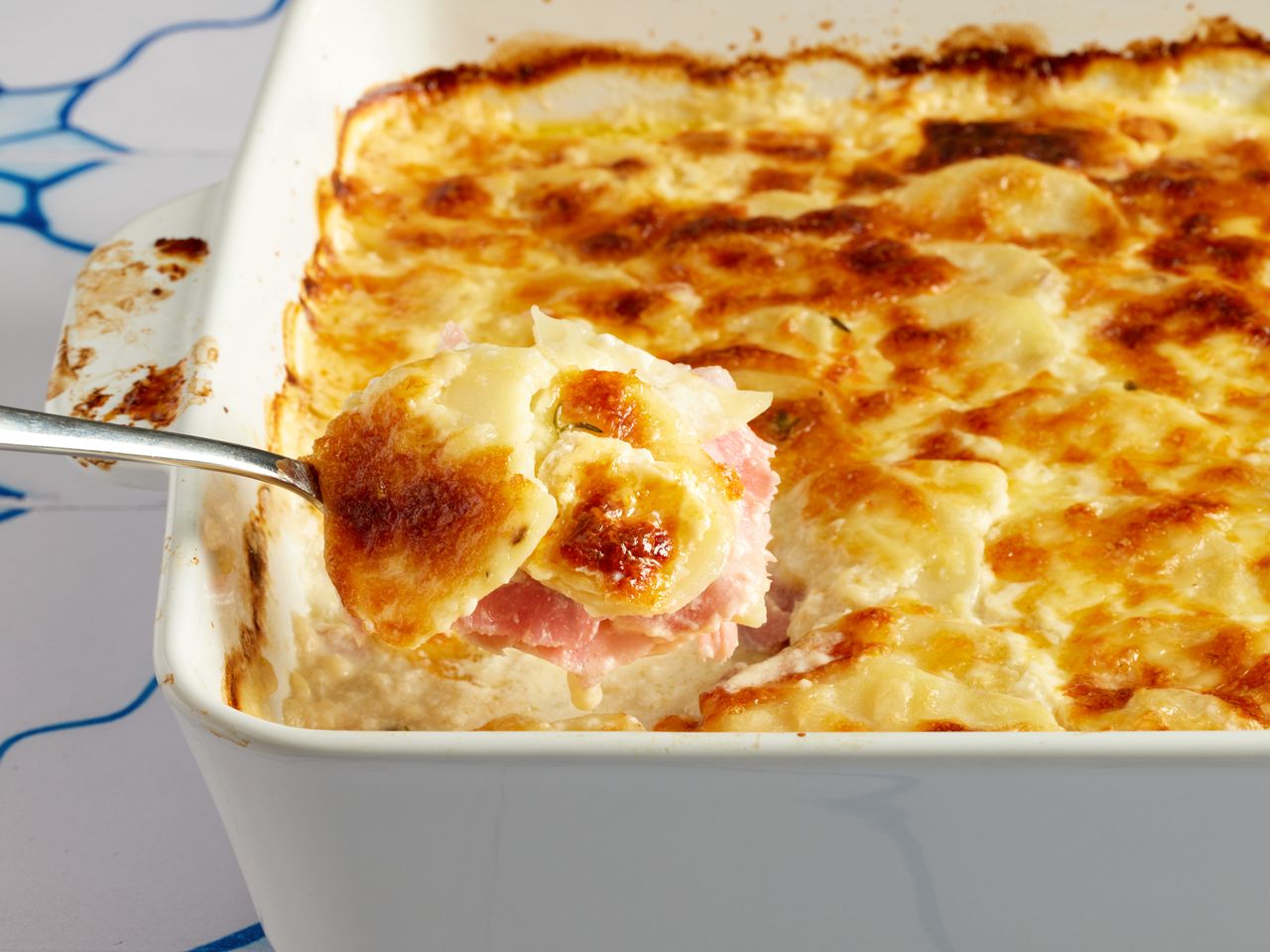 A person removing a scoop of holiday ham casserole from a white casserole dish.