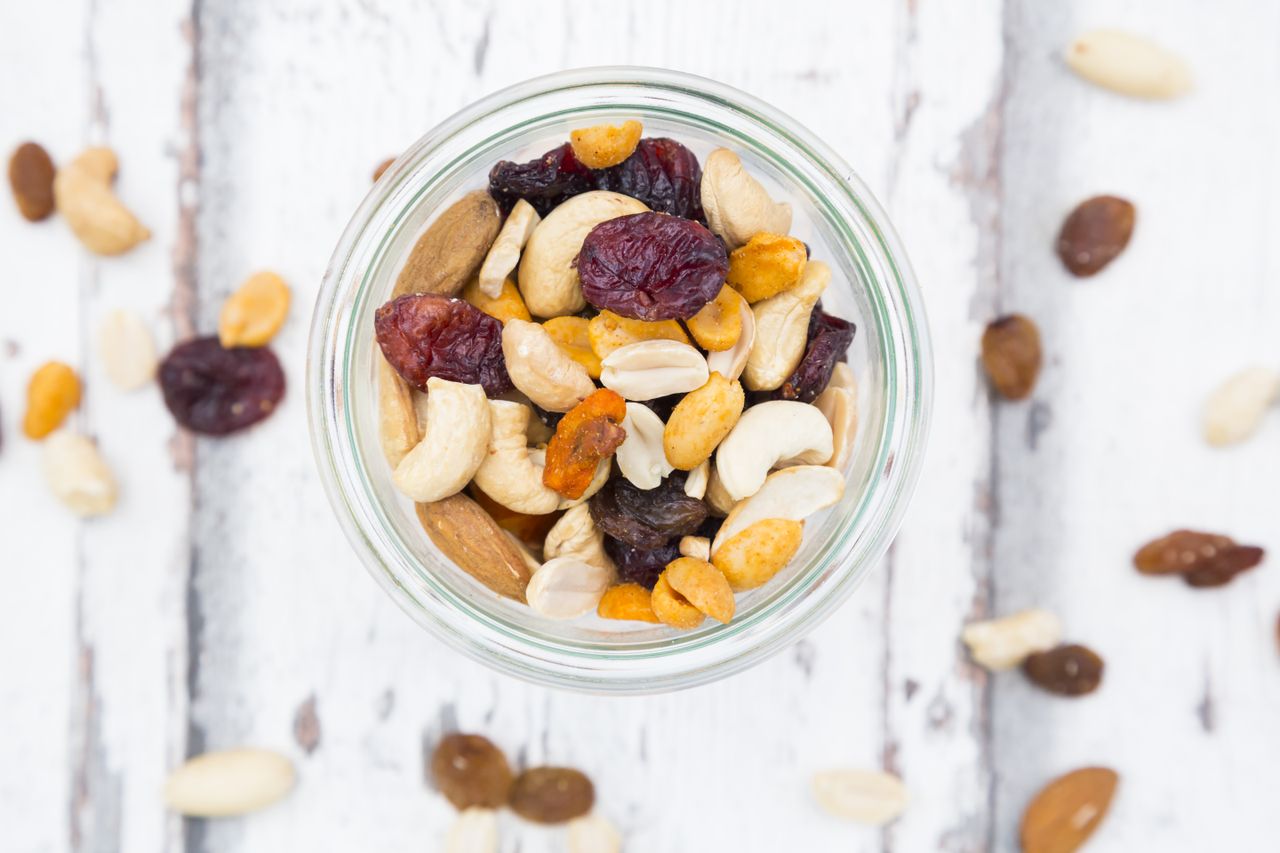A jar filled with mixed nuts and dried fruits surrounded by scattered pieces on a wooden surface