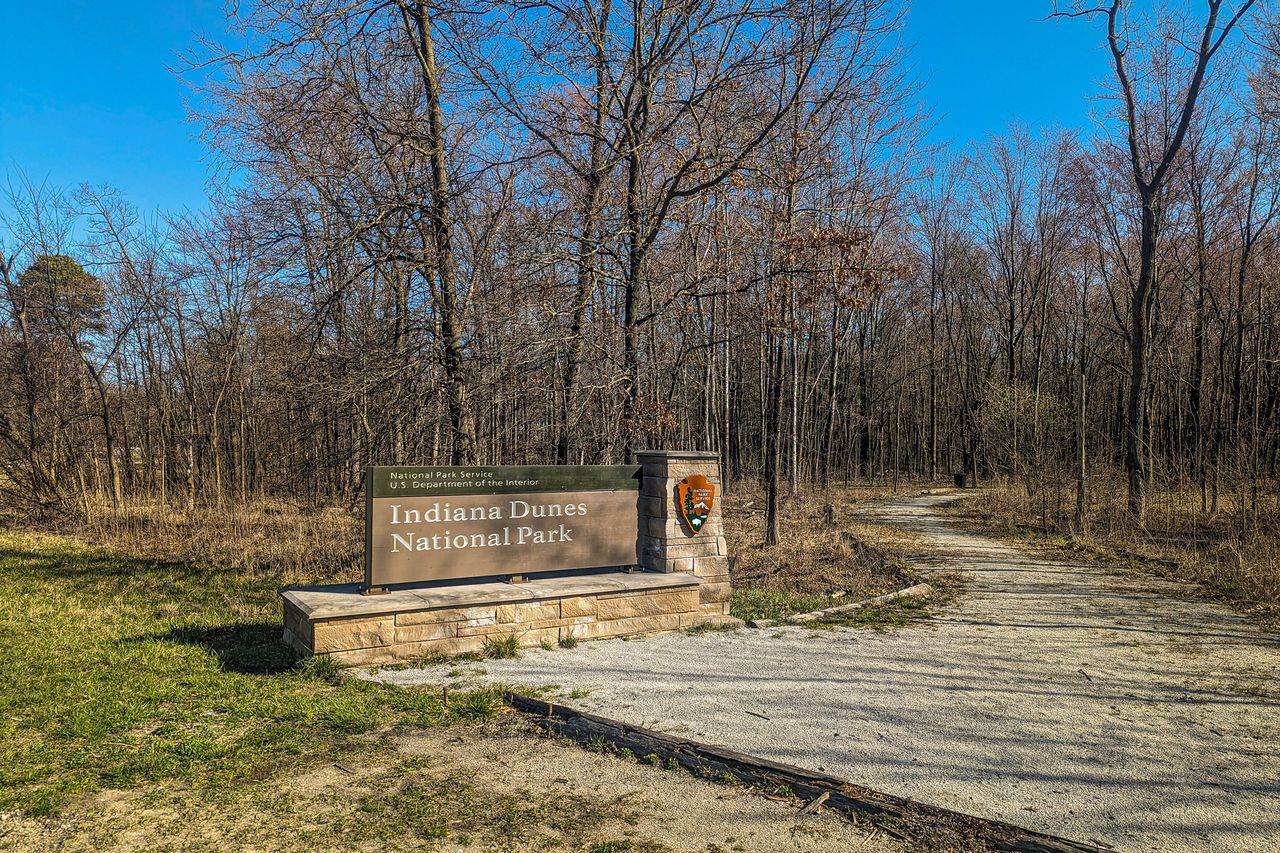 Entrance sign to Indiana Dunes National Park adjacent to a trail leading into the woods surrounded by bare trees and grass