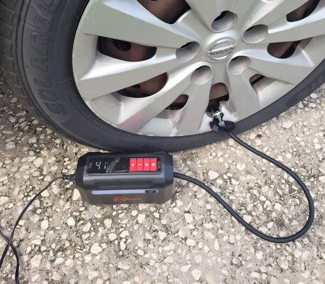 A car tire is being inflated with a portable air compressor displayed on gravel ground, showing a digital pressure reading