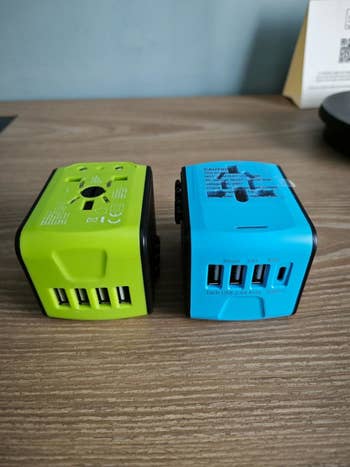 Reviewer's photo of two travel adapters with several USB ports on a wooden table, featuring socket options for various plug types