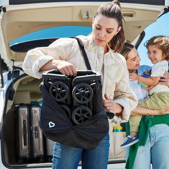 A model holding a folded stroller in a bag near an open car trunk, with another woman holding a child in the background