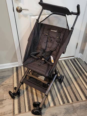 Compact foldable stroller with a padded seat, harness, and sunshade, positioned on a striped rug near a closed door. Ideal for shopping trips