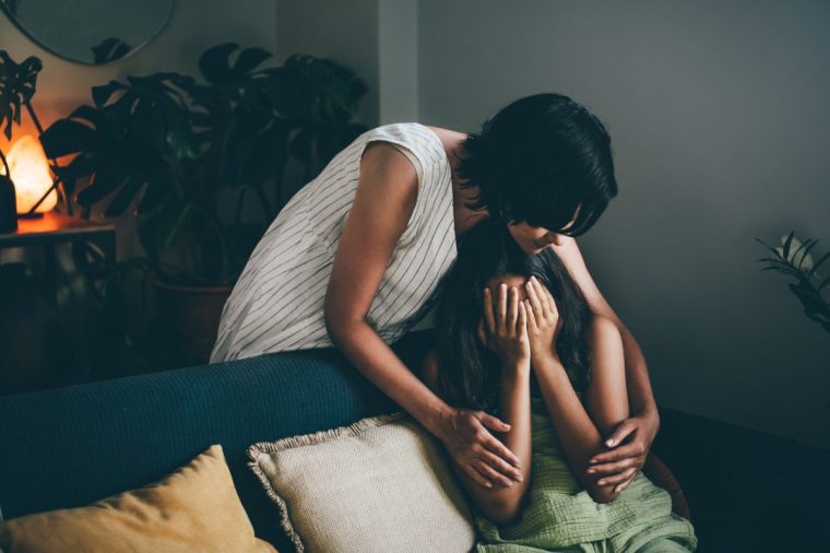 Mother support upset daughter on couch in living room at home.