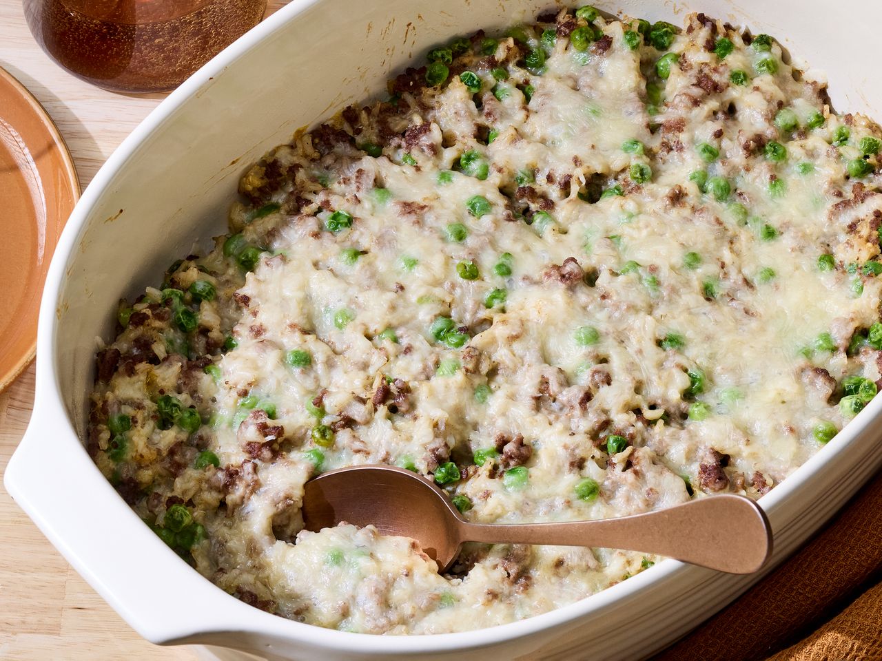 Casserole dish containing ground beef brown rice peas and melted cheese with a serving spoon placed inside
