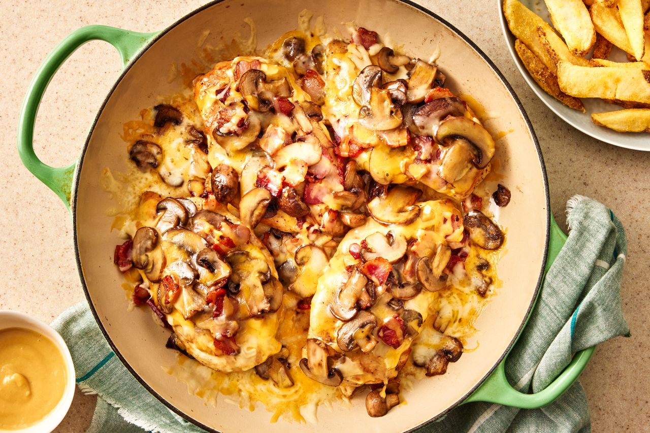 Overhead view of a green pan with four chicken breasts cooked with cheese and mushrooms next to a plate of steak fries and bowl of honey mustard