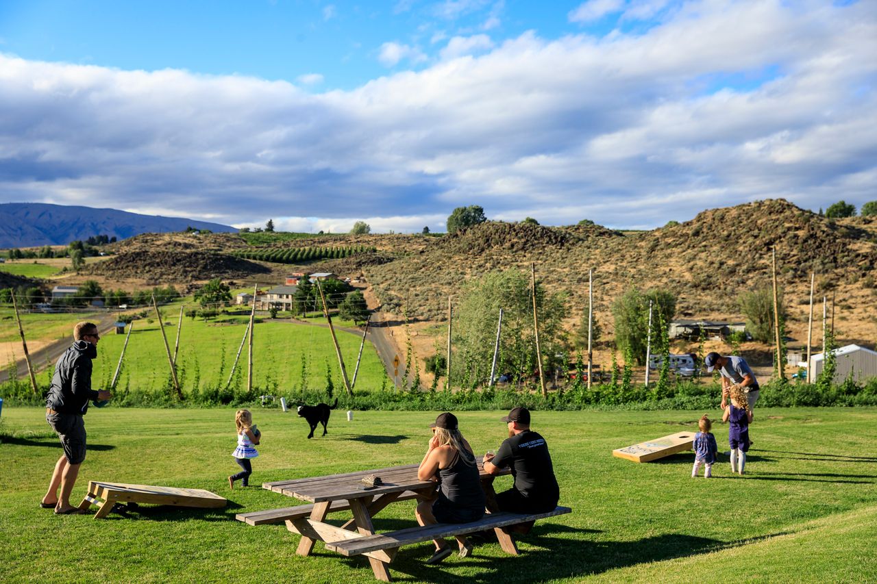 People enjoying a grassy outdoor area sitting on picnic tables with hills and a rural backdrop visible