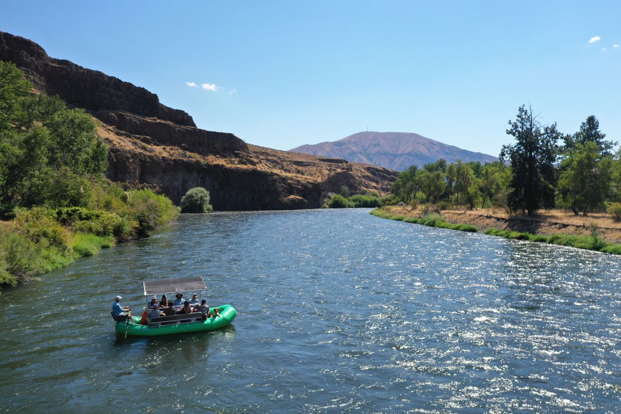 A green raft with people floats down a river surrounded by rocky valleys and sparse trees