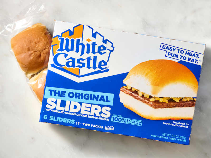 Box of White Castle Original Sliders with two visible sliders in plastic packaging, featuring grilled onions on a signature bun.