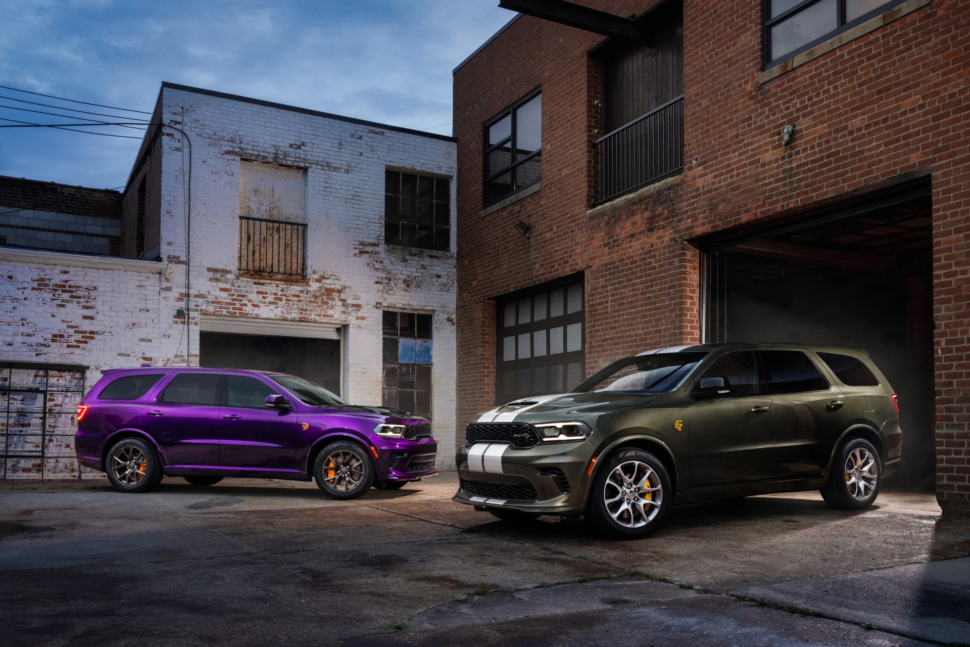 Two Dodge Durango SUVs, one purple and one dark green with white racing stripes, parked in an industrial alley with brick buildings.